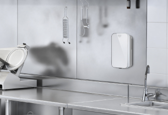 고기 슬라이서와 싱크대 등 다양한 장비를 갖춘 스테인리스 소재의 조리도구들이 있는 주방 벽면에 세스코(CESCO)의 자동 손 세정기 핸드제닉이 설치되어 있습니다.